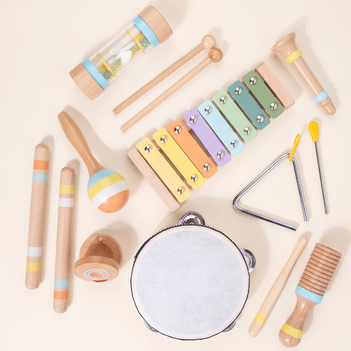 Pastel wooden musical instrument set laid out flat on a cream background: xylophone, tambourine, rain stick, maraca and more